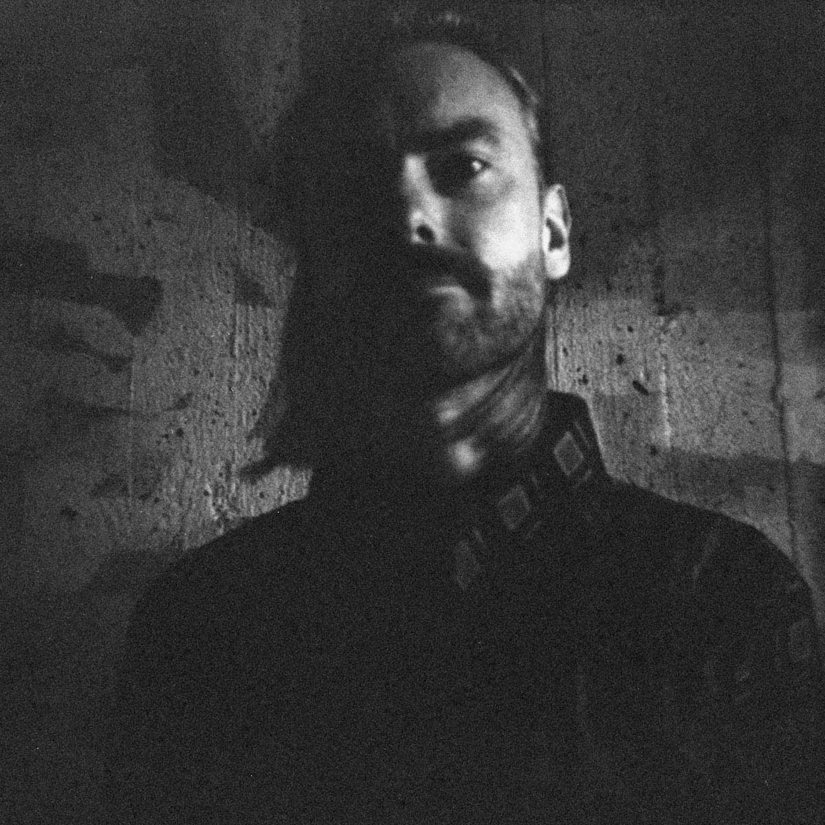 Slightly blurred and dark black and white portrait photo of Karl Vento, with half of his face in shadow. The white man with a black, short full beard stands in front of a textured wall and wears a dark shirt.