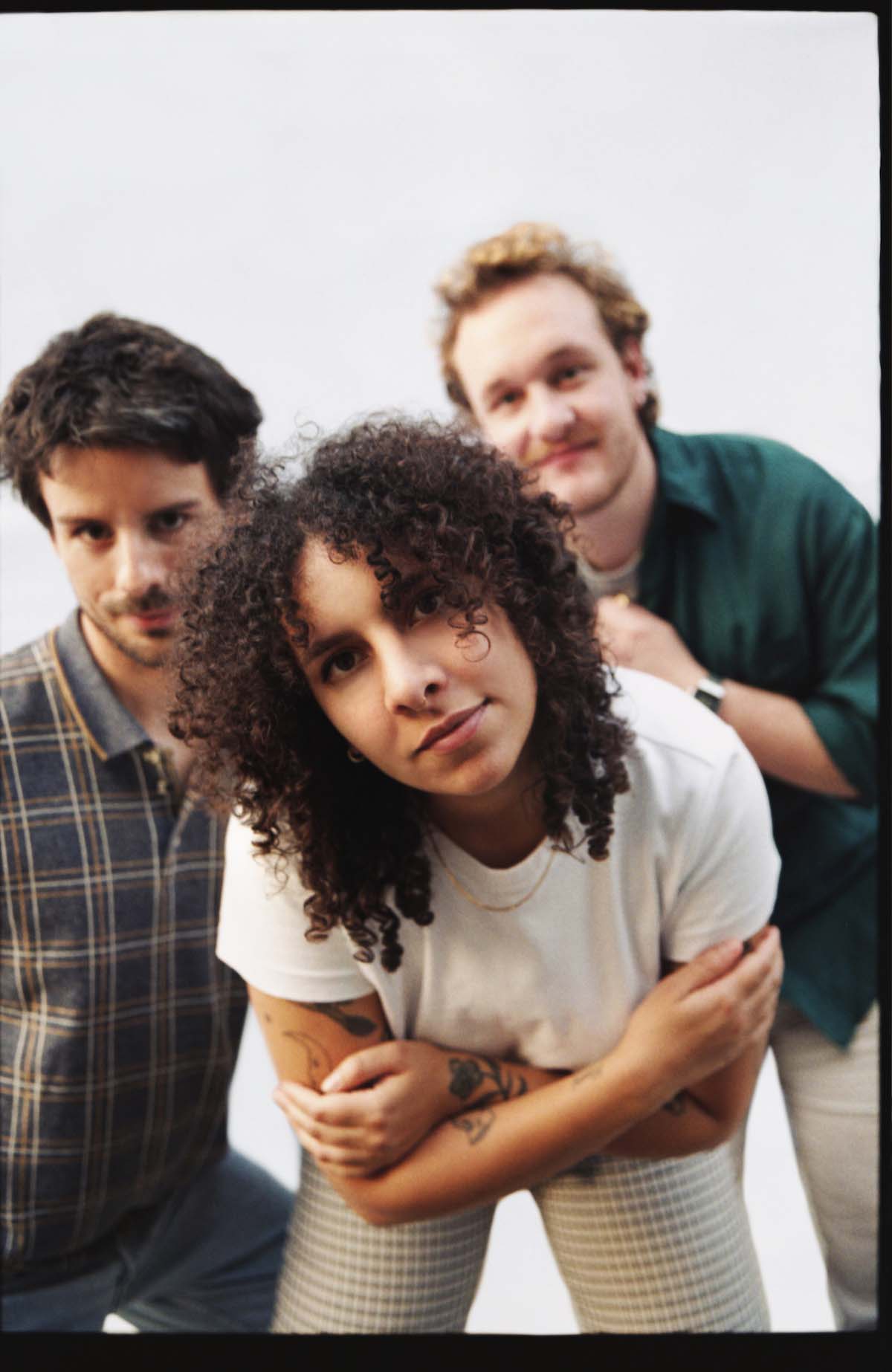 The three members of the band Meagre Martin look kindly into the camera and pose leaning forward. The front woman has dark curly shoulder-length hair, her tattooed arms crossed and is wearing a white T-shirt and light-coloured cloth trousers. Behind her on the left, a white man with a three-day beard, brown hair and a checked polo shirt looks past her. On the right, even further in the background and rather out of focus, a white man with reddish curls, a three-day beard and a dark green shirt can be seen.