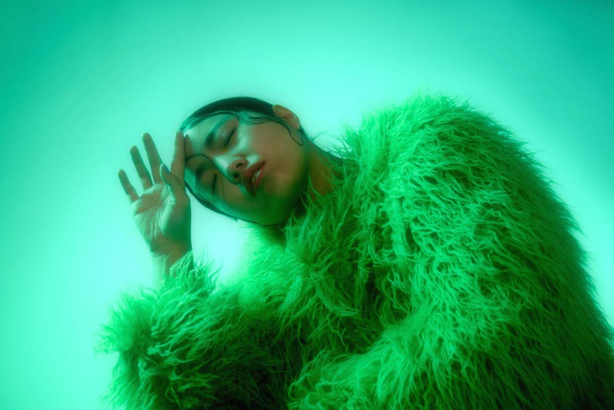 Young, Asian woman is seen in a close-up, she has her eyes closed, her head slightly tilted and touches her forehead with the back of her right hand. mui zyu's black hair is slicked back, single strands fixed to her face. She is wearing a yellow, long-pile, shaggy faux fur jacket. The picture is bathed in green light.
