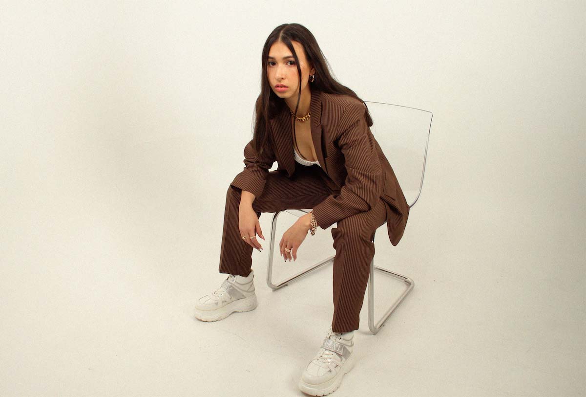 Centrally in the picture, a young woman with dark brown, long hair sits bent forward on a chair with a transparent seat and backrest. She is looking into the camera. The background is completely creamy white. Nashi44 is wearing light make-up, a white top with a plunging neckline, a brown suit and white platform trainers. A strand of her hair falls down her cheek. She props her elbows on her knees, lets her hands hang down with long fingernails and a few gold rings.