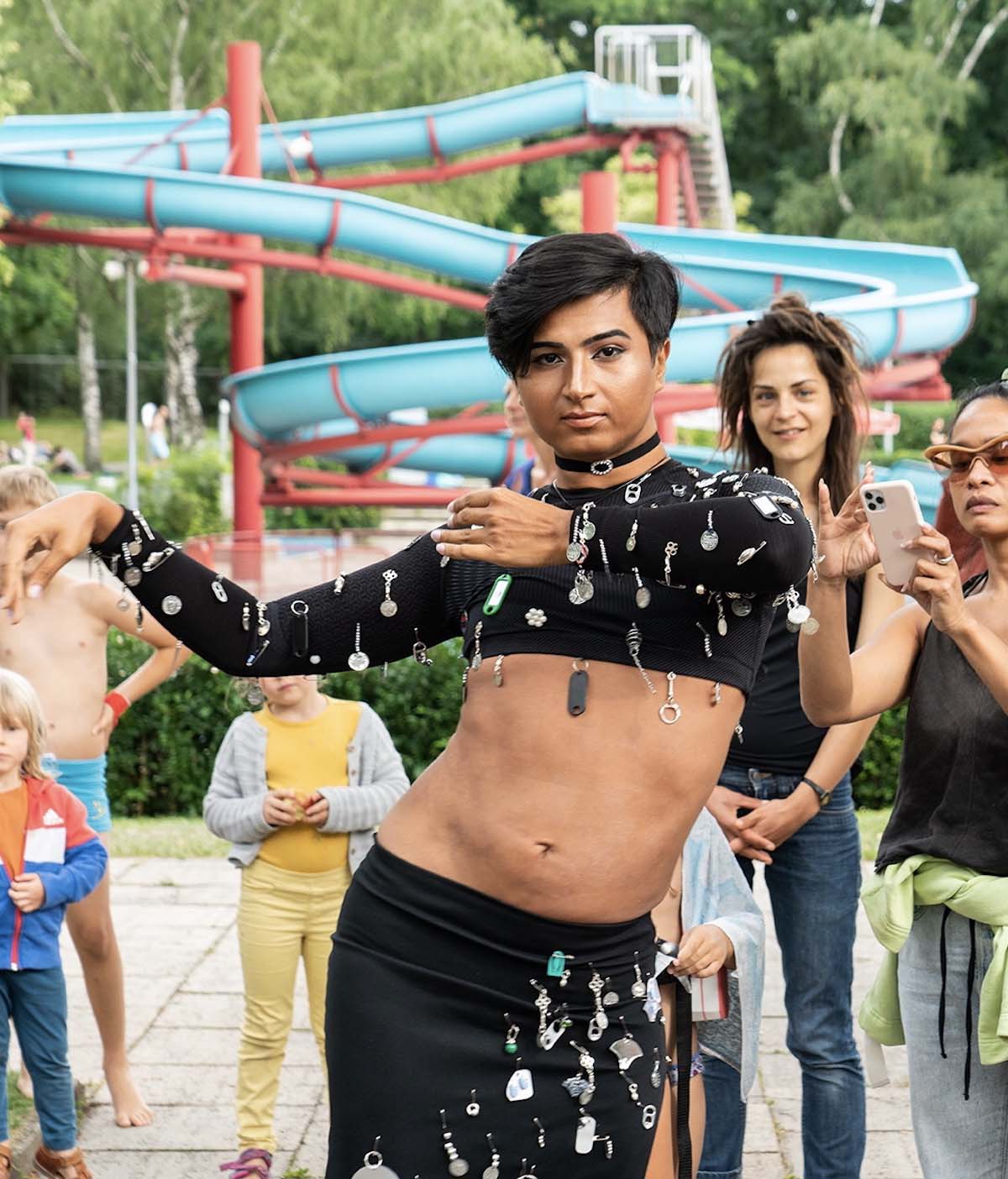 A non-binary Person Of Colour has black temple-length hair, a black choker around her neck and wears a black belly dance costume elaborately draped with various pendants consisting of a low-slung skirt and a long-sleeved very short belly-free top. Prince Emrah is looking at the camera and has assumed a belly dance pose with his hips sticking out and an elegant sideways arm movement. Prince Emrah is standing in an outdoor pool, behind Prince Emrah you can see your long blue slide and onlookers visiting the pool.