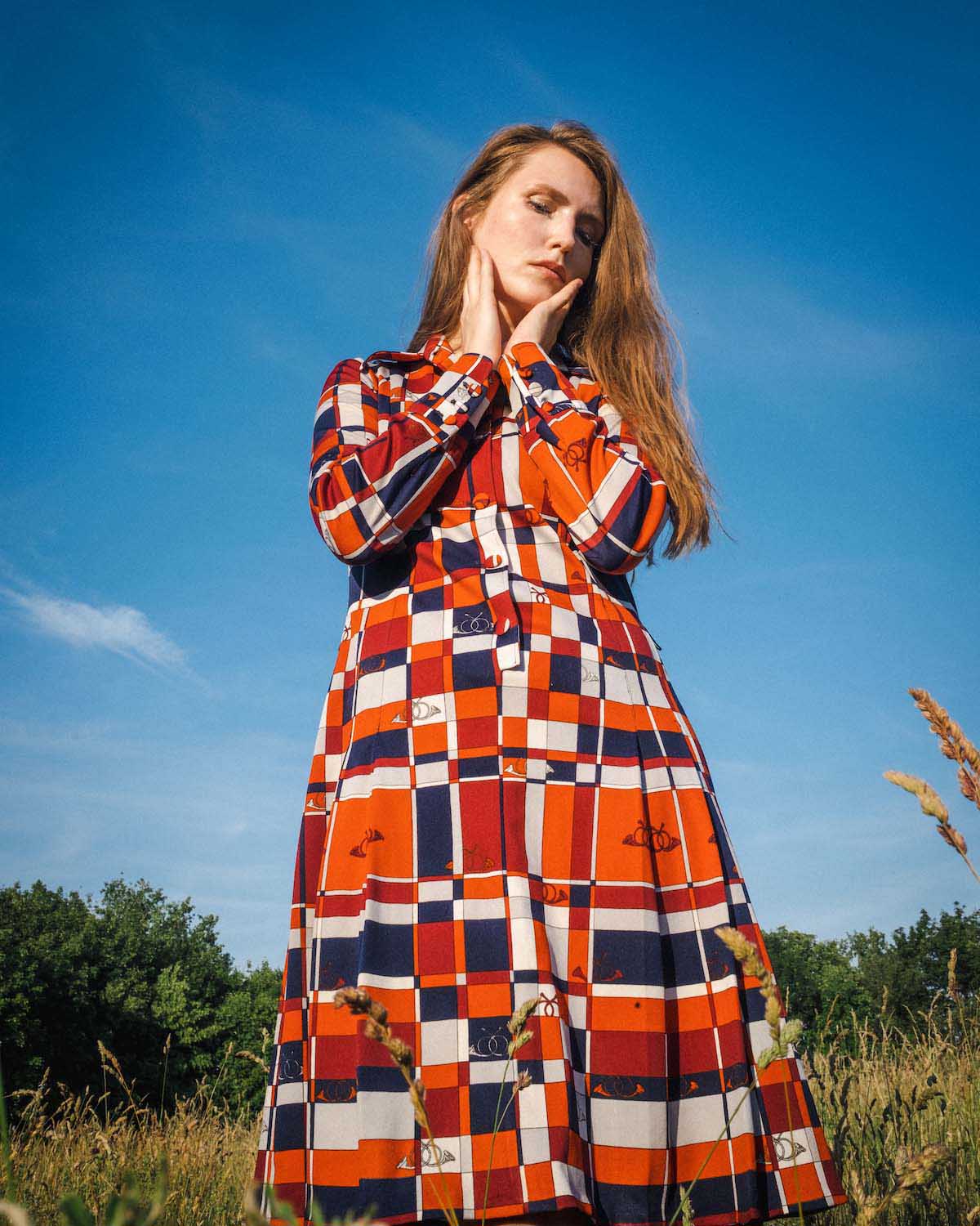 Young, white woman with long red hair stands in a field in a long, long-sleeved dress with a red-white-orange-dark blue tiled pattern. Stella Sommer is photographed from below, so that mainly blue sky can be seen behind her and some trees at the bottom of the picture. She puts both hands on her chin and cheeks and looks diagonally downwards in a pensive way.