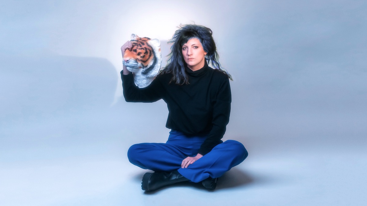 Le Tigre Bleu, a young white woman with long dark hair with a blue shimmer, sits cross-legged on the floor against a white background. She wears a black long-sleeved blouse, blue trousers and heavy black boots. In her right hand she holds a rubber tiger mask at face level next to her.