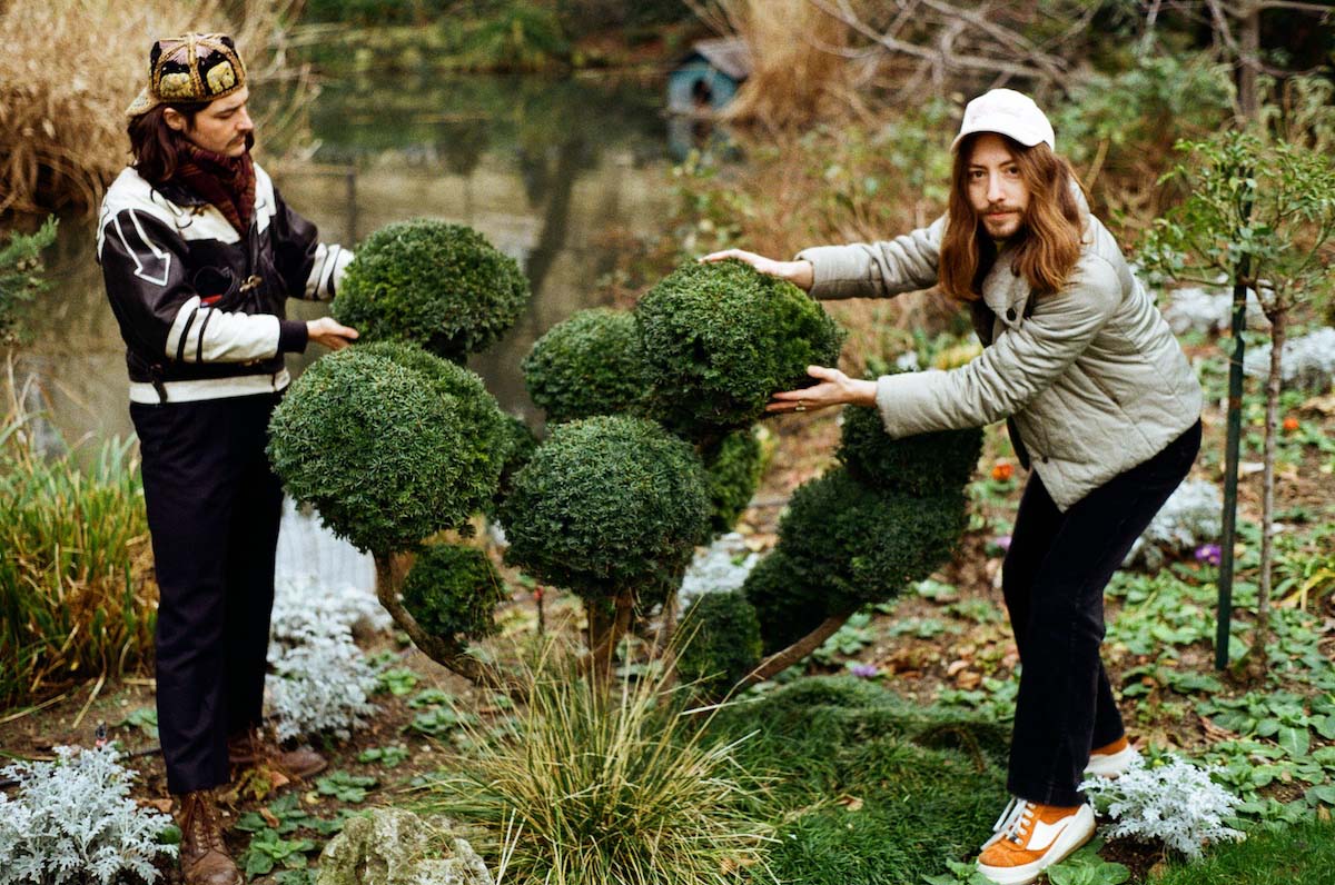 Two white young men with long brown hair stand respectively to the left and right of a bush whose leaves form large balls at the end of the branches. The two members of the band Walter Astral touch these balls as if presenting the bush, which is in the middle of a park with a pond in the background. The one on the right looks into the camera, wearing a white baseball cap, moustache and chin beard, a grey quilted jacket, black trousers and orange and white trainers. The one on the left looks reverently at the bush, he has a cap on with the shield facing backwards, wears a moustache, a black and white biker jacket, black trousers and brown shoes.