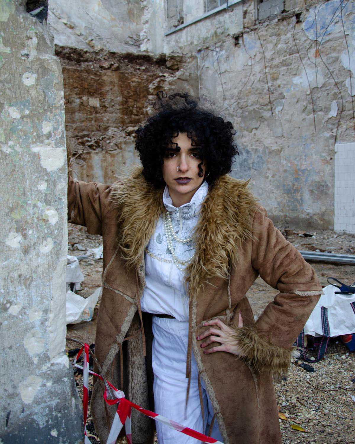 Young woman with black, chin-length curly hair stands between the walls of a building in ruins, in front of her some red and white tape. Rasha Nahas looks into the camera, leans against the wall of the house with one arm and rests the other on her hip. She wears a white tracksuit, white pearl necklaces and a brown lined suede coat with fake fur appliqués on the collar and sleeves. Her lips are darkly made up, she wears a nostril ring.