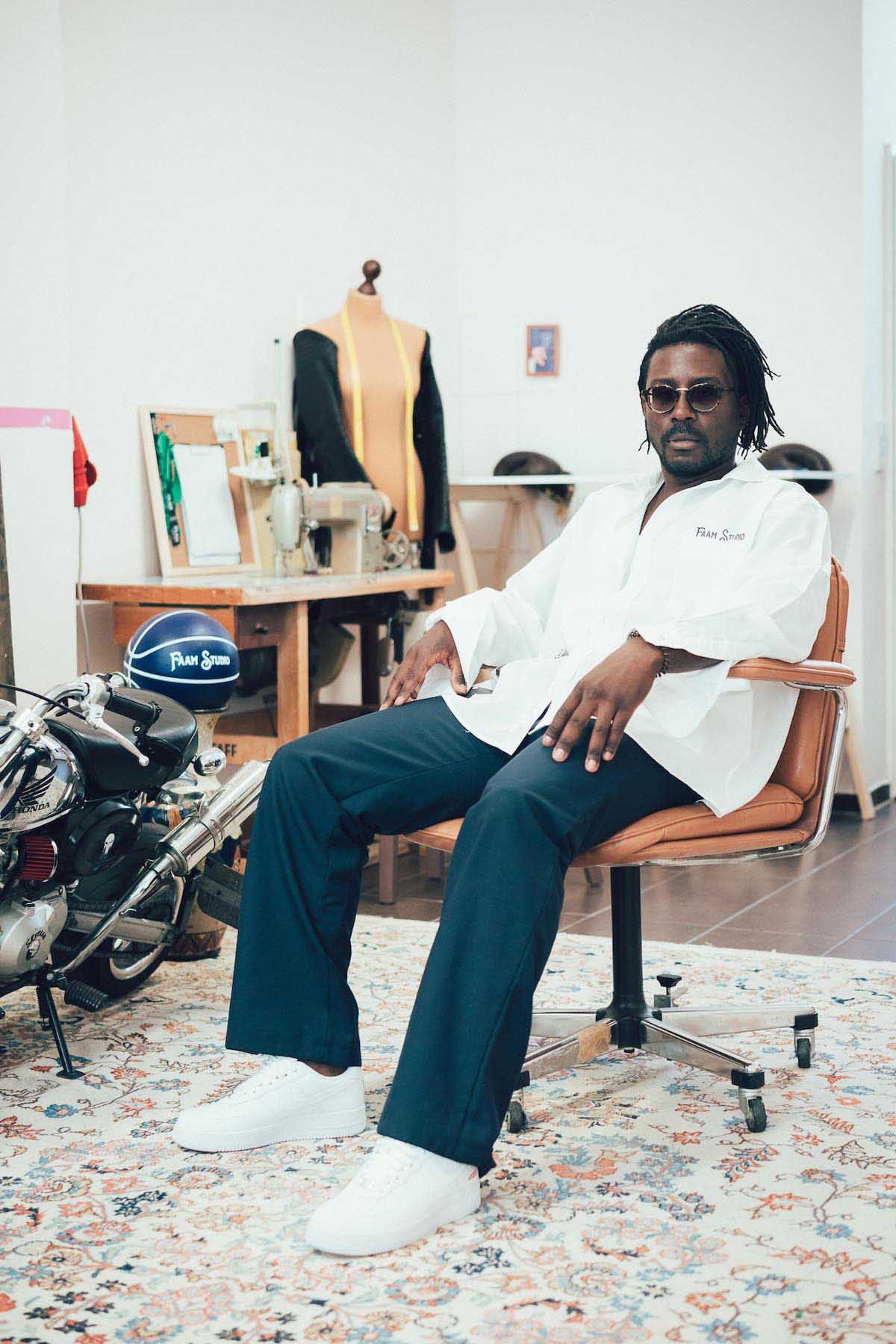 Stéphane Moun alias FAAM, Black person with thin, neck-length dreadlocks sits casually on an orange chair with wheels, which stands on a large carpet. He wears a moustache and chin beard, a white oversized shirt, dark blue trousers and white trainers. He rests his arms loosely on the backrests and looks at the camera. Behind him, a tailor's dummy with a tape measure around its neck and a sewing machine can be seen. To his left there is also a motorbike for decoration.