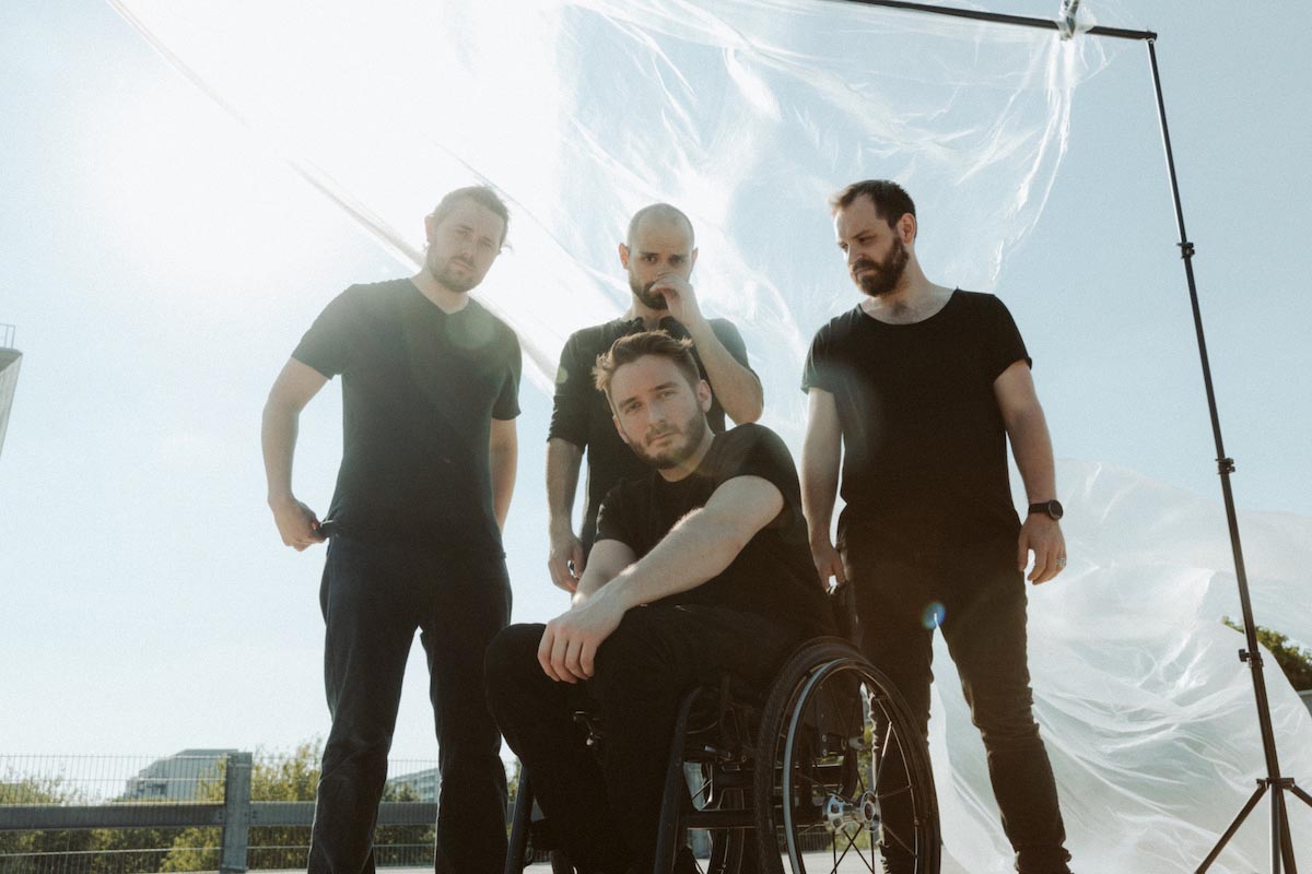 Four men, all dressed in black T-shirts and black trousers and with dark beards, pose outdoors in front of a transparent plastic sheet on a stand that is blowing in the wind. The front man of the band Fheels sits in a wheelchair at the front, has his arm resting on his leg and is looking at the camera; behind him are the other three members. The one on the left in the picture is adjusting his T-shirt at the back, the one next to him with a bald head is grabbing his nose, both are also looking at the camera. The fourth looks grimly to the left. A fence and residential blocks can be glimpsed in the background.