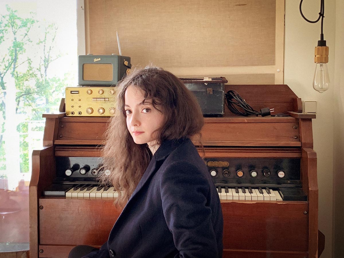 White, young woman with long curly brown hair sits turned in towards the camera in front of a brown, retro home organ with vintage radios on it. To the right of Gloria de Oliveira hangs an equally vintage light bulb. Gloria can be seen up to her waist and is wearing a heavy dark blue jacket, with her hair tucked into the collar on the right.