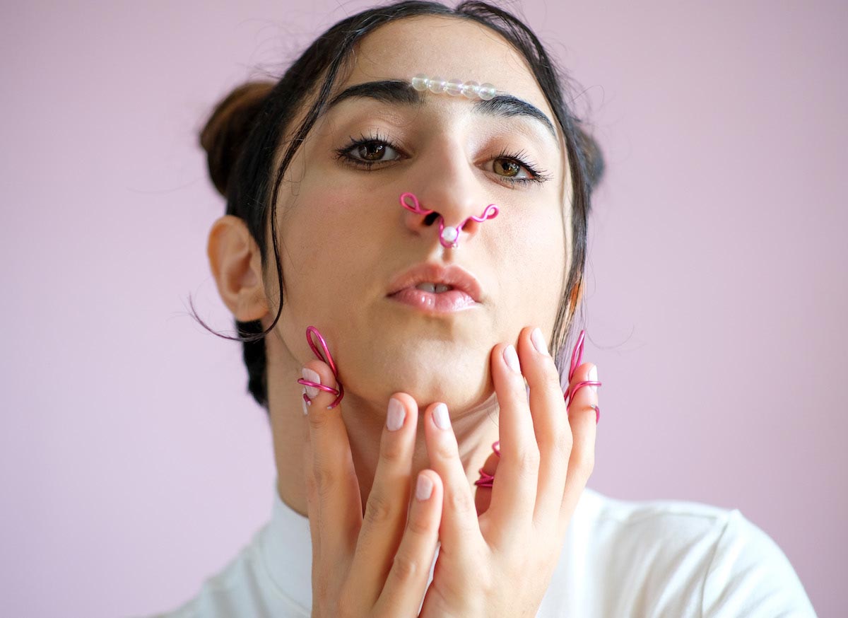 Portrait photo of a young woman with black hair tied into space buns. MADANII is looking directly into the camera and has her lips slightly parted. Five transparent pearls are attached in a row between her black eyebrows. She touches her chin delicately with both hands. In her nostrils and on two fingers she has attached pink curved wire formations.