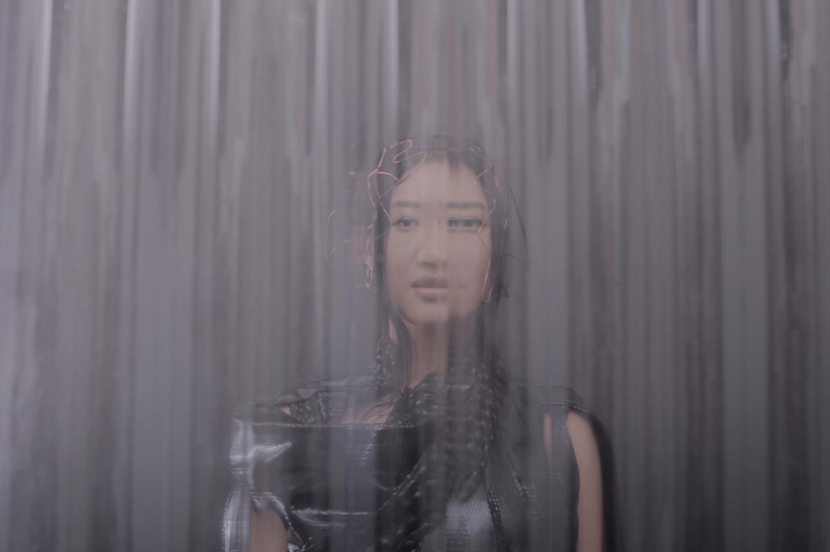 Young Asian woman with black hair can be seen up to below her shoulders looking at the camera. Portrait XO is wearing a black, leather-looking warrior top, with copper-coloured wires entwined over her head. The image is distorted as if looking through a waterfall.