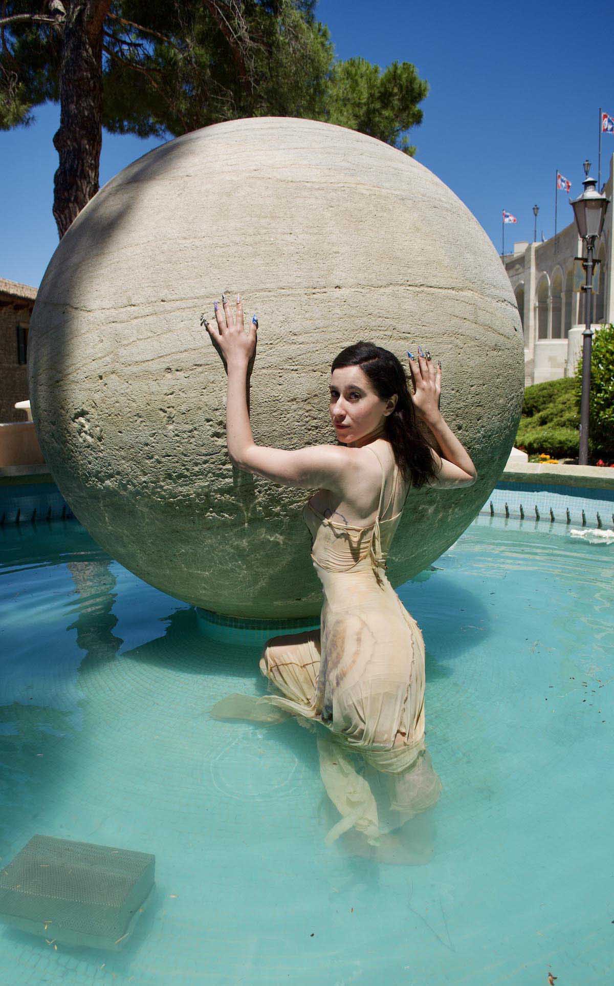 Young white woman with long dark hair wears a beige, long, fine spaghetti strap dress. Selin Davasse stands with her back to the camera in a fountain whose water looks light blue, in front of a stone sphere, also beige, which towers over the artist. She touches the sphere with both hands, on which she wears long colourful fingernails, and looks over her shoulder into the camera. In the background is a blue sky, a pine tree and a southern building.