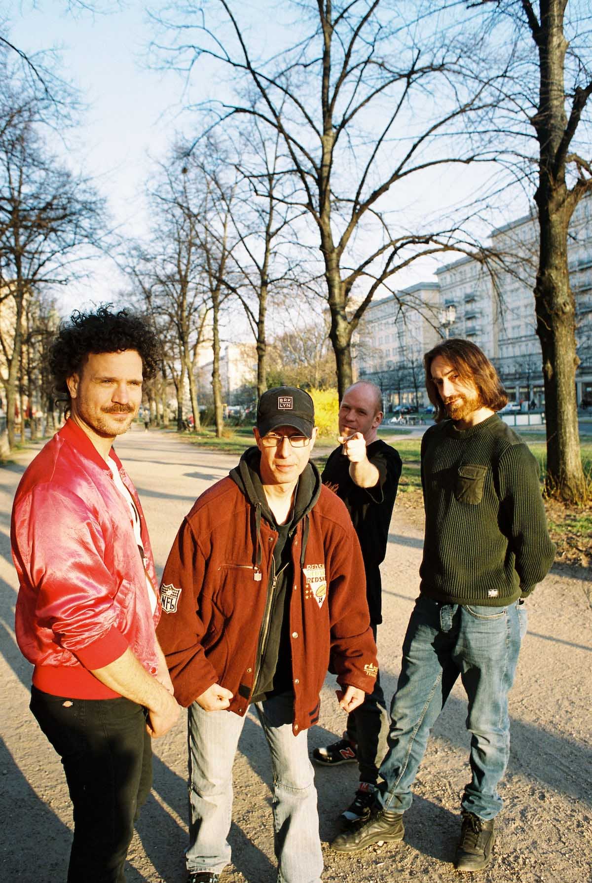 The members of the band Wellen.Brecher - four white men - are standing next to each other on a wide dirt footpath avenue, the trees are bare, multi-storey apartment buildings can be seen in the background. The one on the left has longer black curly hair, a moustache and wears a shimmering red blouson and black jeans. The one next to him has glasses and a black baseball cap on, wears a black hoodie, light jeans and a dark red NFL jacket. He looks into the camera. The one slightly behind him is pointing his finger at the camera, he has little hair and is dressed all in black. The one on the far right has chin-length brown hair, a full beard, a dark green coarse jumper, blue jeans and black shoes.
