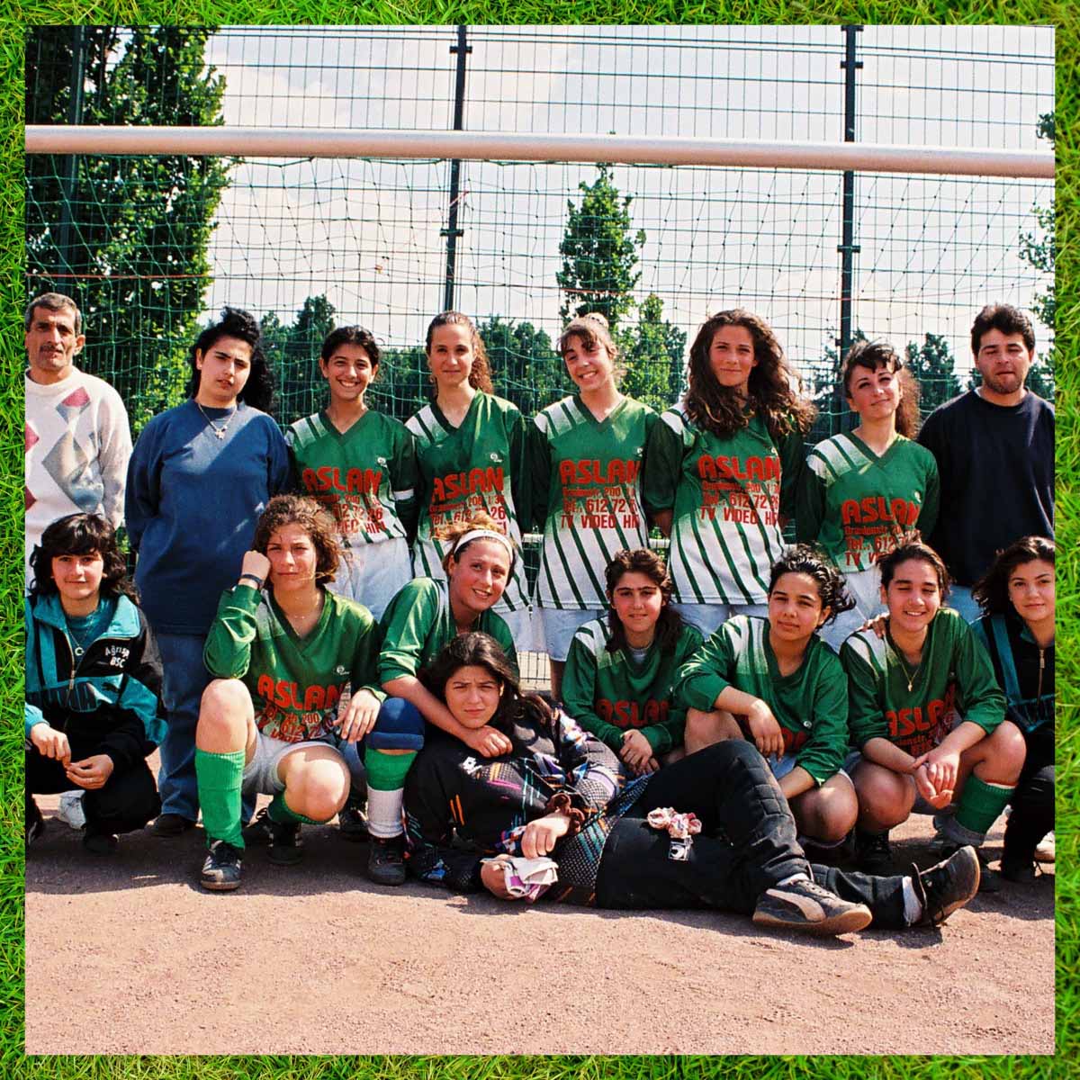 Team picture of the BSC Agrisport girls in front of a football goal. The back row stands, framed by two men, the front row kneels and the goalkeeper lies in front of the front row and is embraced by a teammate. Almost everyone is wearing the green and white jerseys of their club and looking at the camera. There is a frame around the picture showing green football turf symbolising the football focus of Pop-Kultur.