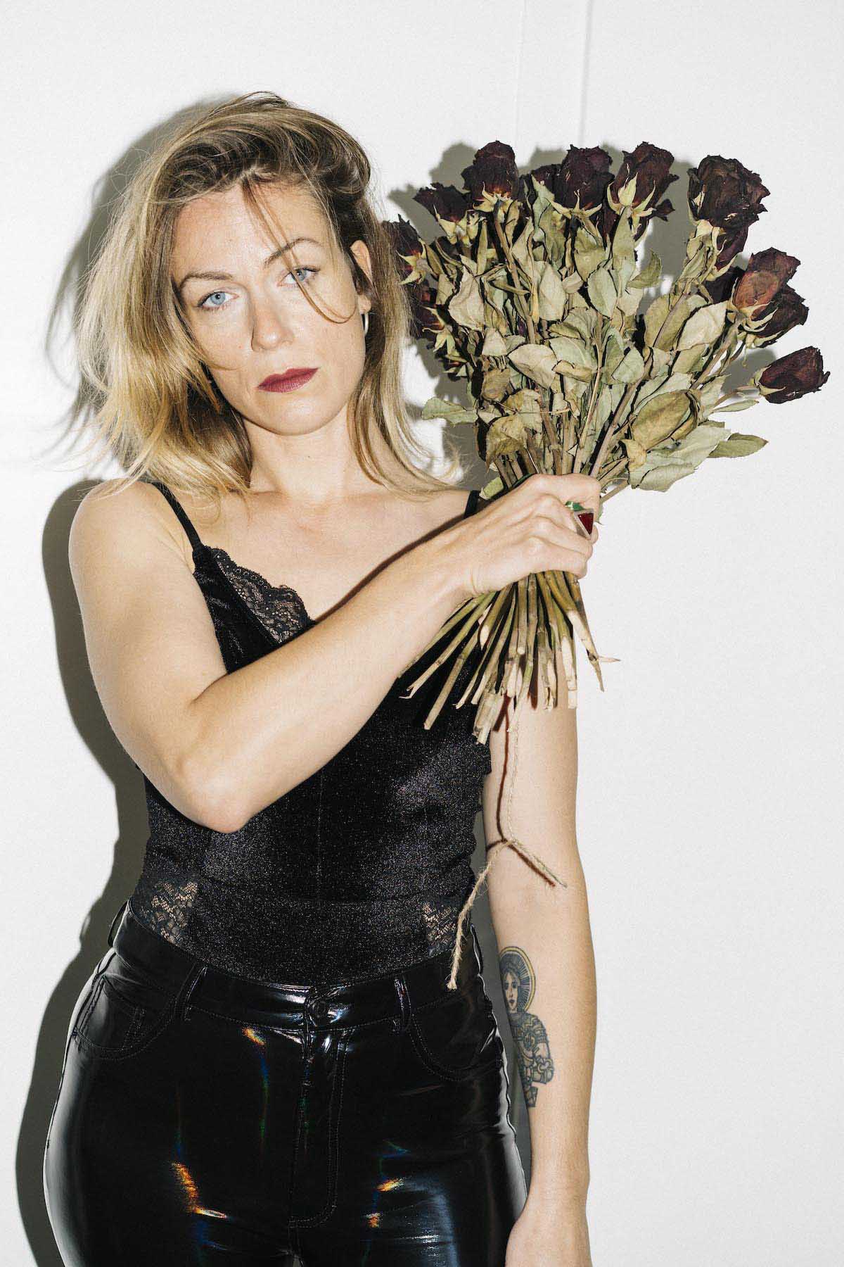 Blonde, white woman with shoulder-length hair, a black tank top with ruffles and black vinyl trousers looks into the camera. Charlotte Brandi has piercing blue eyes, her lips are red. A tattoo can be seen on her forearm. With her right hand, she holds a withered bouquet of roses next to her face towards her left shoulder. The background is white.