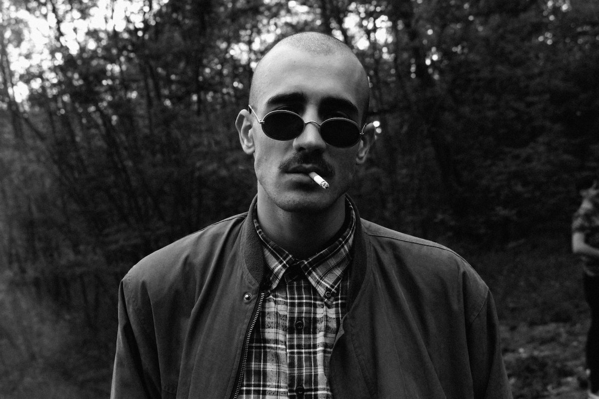 Half close-up black and white photo of a young white man. Morwan is standing in front of a bush, bald, wearing sunglasses with oval lenses, a checked shirt with a blouson, a moustache and a half-burnt cigarette in his mouth. He looks serious at the camera.