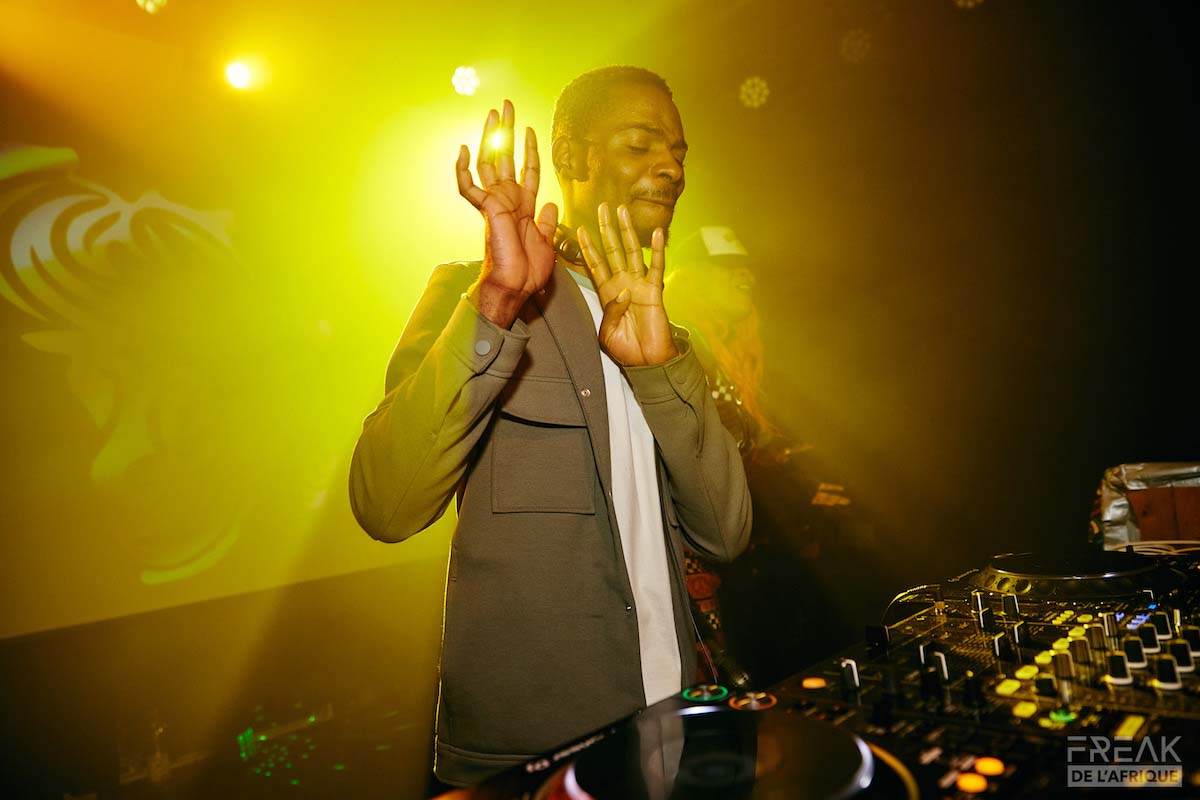 Male Person of Colour with short black hair and moustache stands behind a console. DJ Jeff looks down at the controls and dynamically moves his hands upwards with fingers extended in front of his face. You can see he is moving to the music and has his lower lip pulled in. He is wearing a khaki jacket with a white T-shirt peeking out from underneath. Behind him, another person can be glimpsed in the glaring yellow stage light.