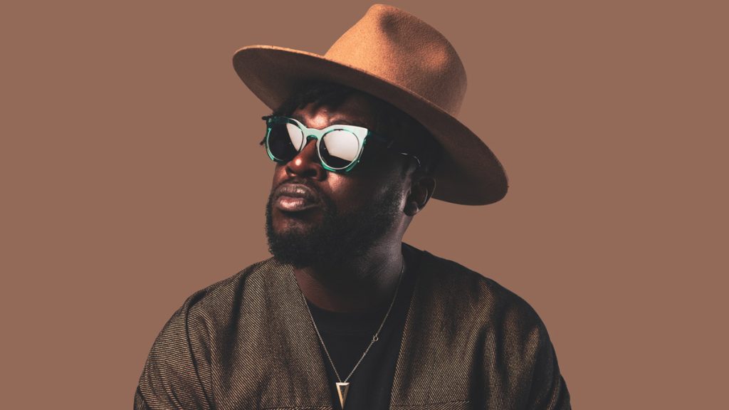 A Black male person can be seen in a monochrome close-up against a brown background up to his shoulders. Blinky Bill is looking to the top left of the picture. He is wearing a black beard, green sunglasses, a brown hat and a necklace with a golden triangle pendant.