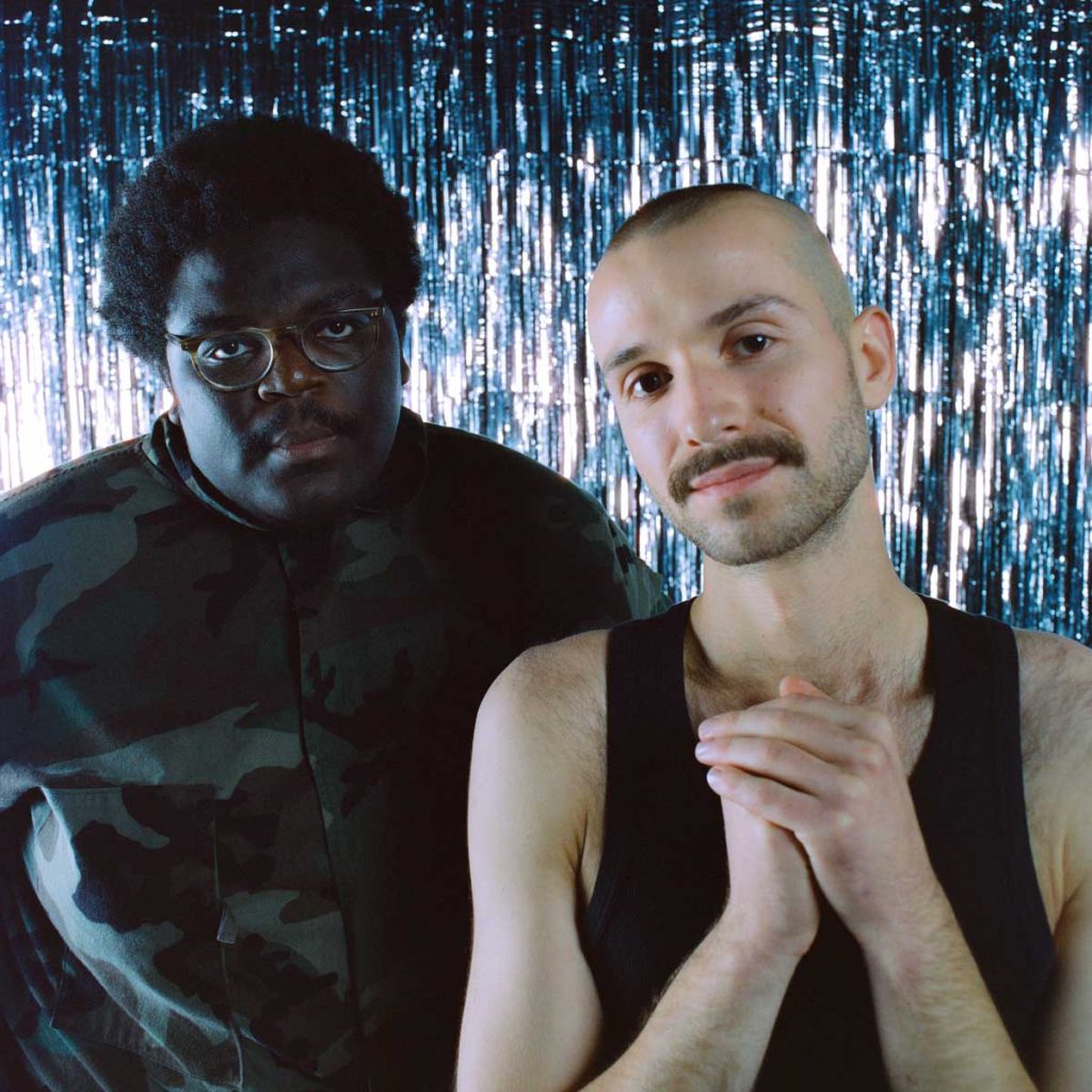 The duo Blumengarten stand in front of a glittering silver wall of tinsel and look into the camera. Rayan on the left, a Black man with a moustache and glasses, is wearing a dark shirt with a camouflage pattern, Sammy next to him, a white man with a moustache and bald head, is wearing a black tank top and clasps his hands together in front of his chest.