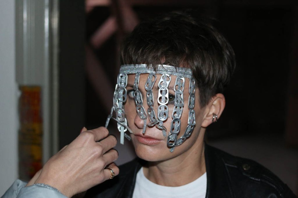 Close-up of a white woman with short brown hair looking at the camera through a veil of can tabs in front of her face. It is Cheap Wedding. The hand of another person can be seen, apparently fastening the tabs.