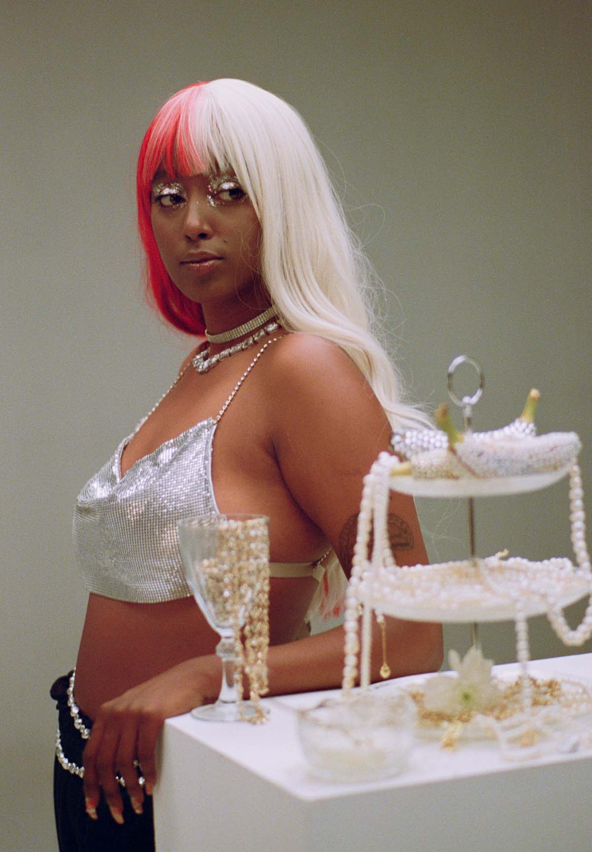A black woman with a blonde-pink long wig and a glittering silver crop top with spaghetti straps leans casually against a white bar table, on which stands an etagere full of pearl necklaces and a glass with gold chains. Eden Derso's eyes are highlighted with glittering silver make-up.