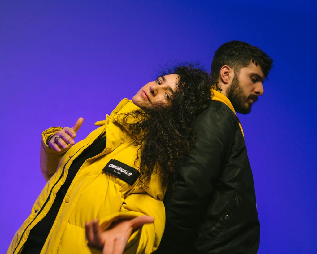 The duo EsRap can be seen against a blue background. The woman, with long curly brown hair, stands on the left, wearing a yellow puffer jacket with a black top underneath, she points to the camera with a casual hand gesture and leans against the man. He has black hair, a full beard and is wearing a black jacket under which a yellow jumper can be seen.