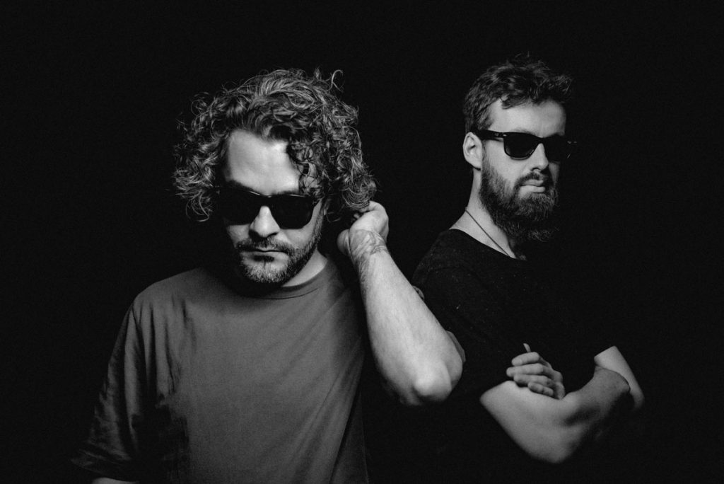 Black and white image against a black background of two men, both wearing sunglasses, monochrome T-shirts and beards - the band Esfand. The man on the left looks down and reaches into his curly, half-length hair. The one on the right stands to the side, wears his hair short, has his arms crossed and looks blankly at the camera.
