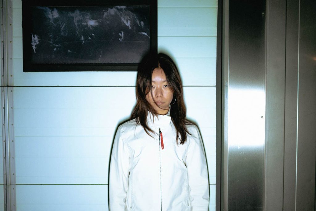 A woman with long dark hair falling into her face stands in front of a white wall with a dark picture hanging on it. EuroEyez wears a white jacket and looks seriously into the camera.