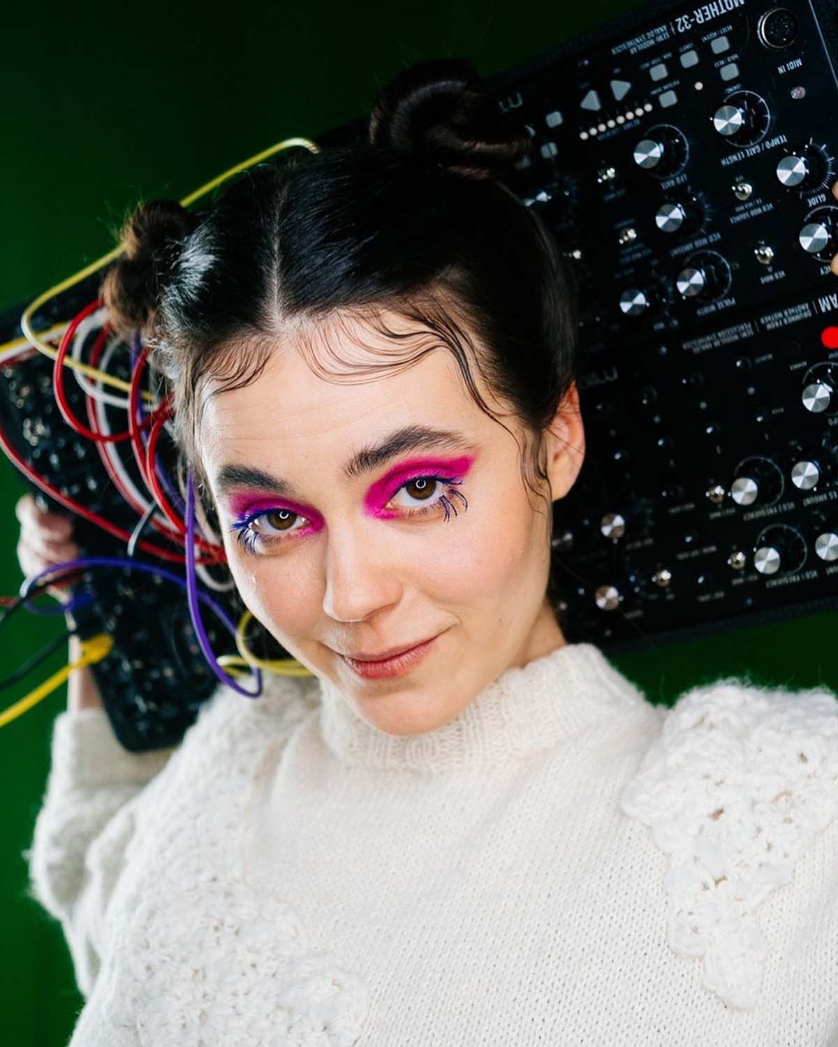 Close-up of a white woman with heavily pink-rimmed eyes, her dark hair parted in the centre and pinned up in double buns. Evija Vēbere smiles slightly into the camera. She is wearing a white woollen jumper and is holding a mixer with colourful cables plugged into it behind her head.