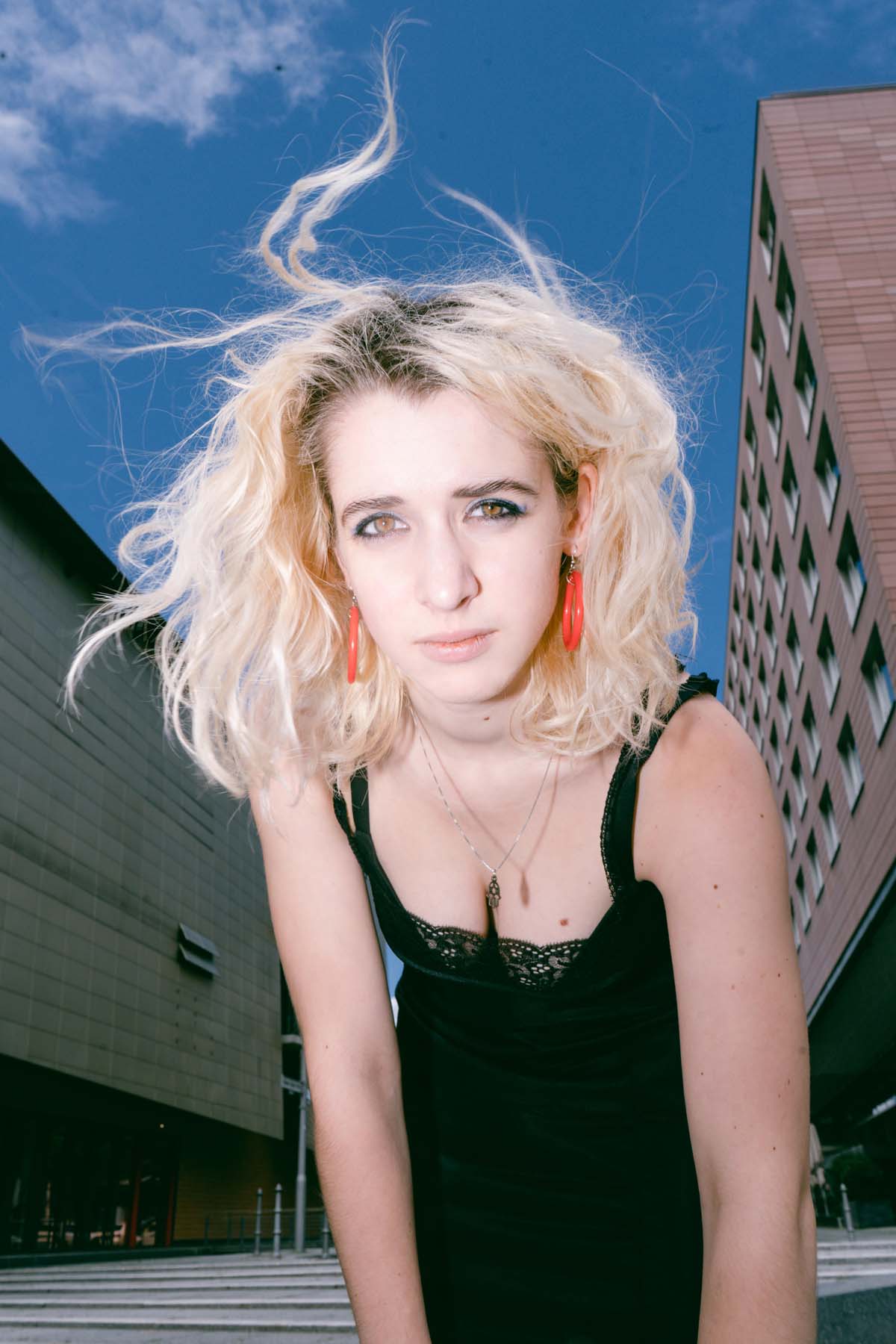 White woman with blonde hair leans forward with a serious expression. Fee Aviv has blonde hair that is blown by the wind. She is wearing a black spaghetti strap dress and orange-coloured hoop earrings. Behind her is a city backdrop with multi-storey buildings and a blue sky.