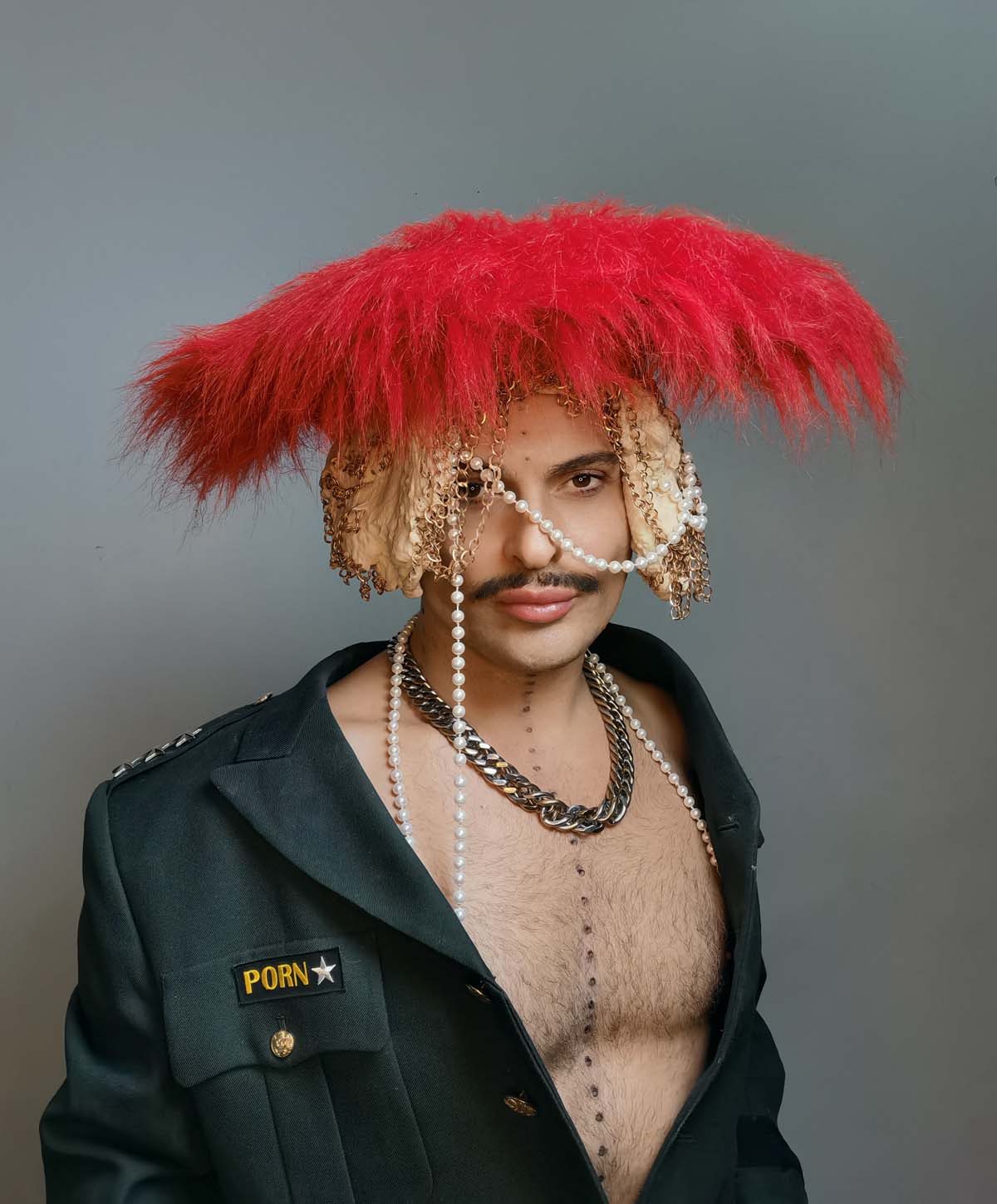 White male can be seen up to the waist against a grey background. Ivo Dimchev is wearing a dark grey uniform jacket with "PORN" written on the breast pocket. His chest is bare. He wears a moustache, a heavy link chain around his neck and a mushroom-shaped headdress made of tulle, gold chains and a wide, bright red top made of long plush hair. A white pearl necklace is draped around his head and shoulders.