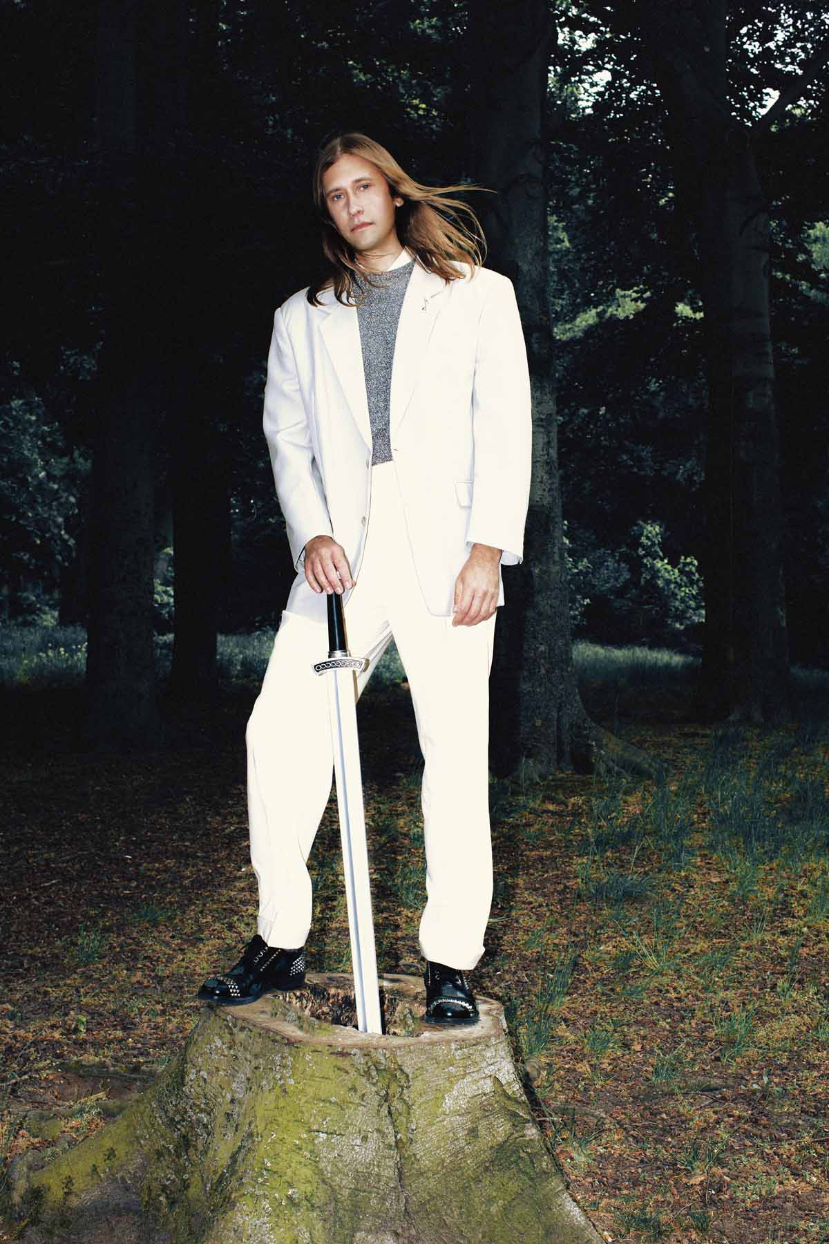 White man with long light brown hair stands in a forest on a tree stump with a sword stuck in it. Jaakko Eino Kalevi is wearing a white suit, black shoes and a grey top under his jacket. His hair is slightly blown by the wind, he has one hand loosely on the hilt of the sword and looks into the camera.