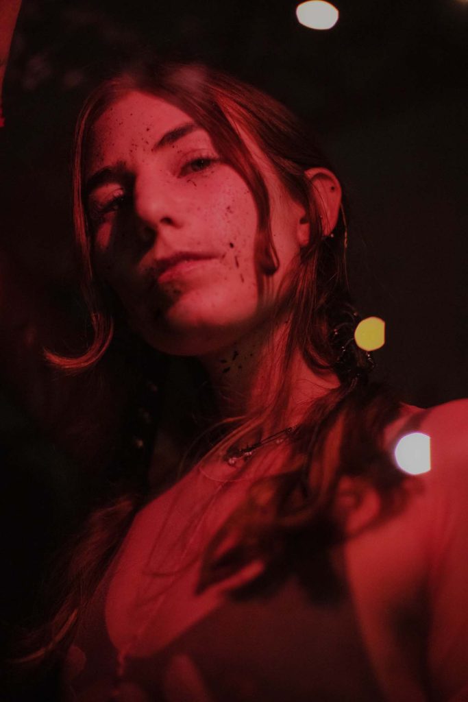 A slightly blurred portrait of James K, a white young woman with freckle-like black dots on her face and brown hair, strands of which fall into her face, photographed from below at an angle and bathed in red light.
