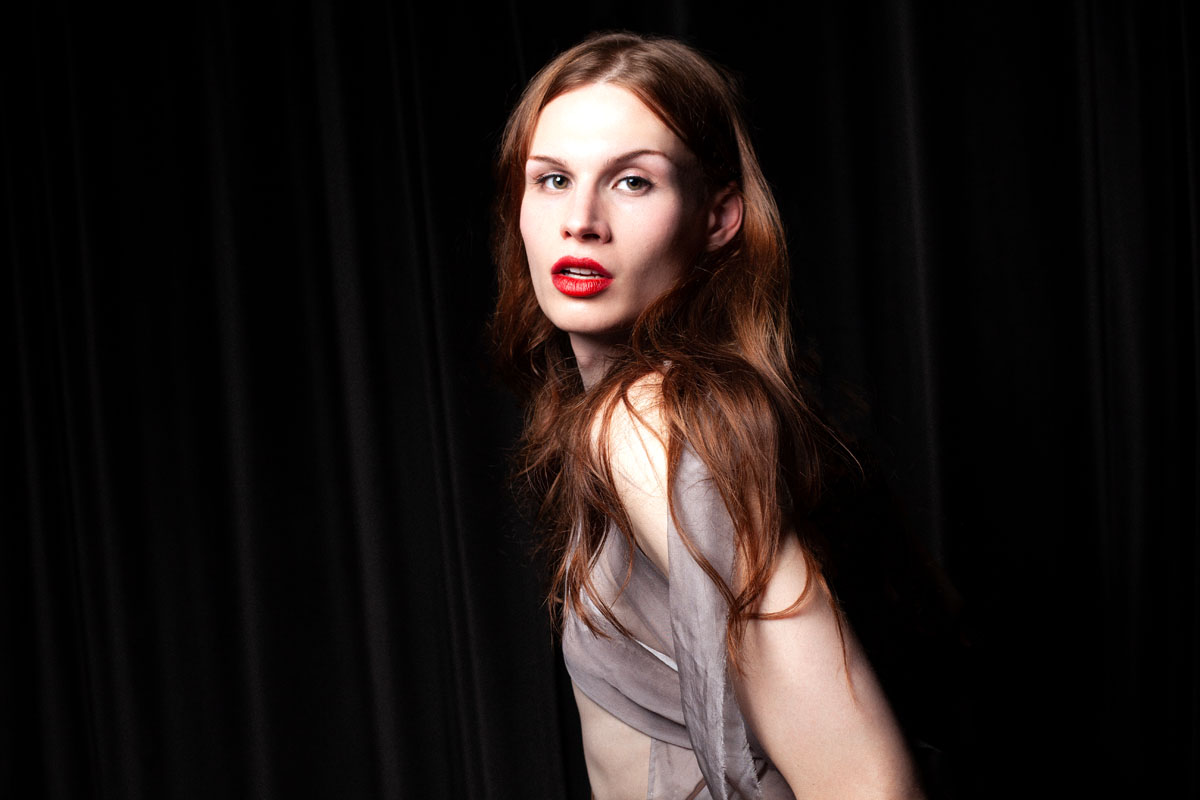 White female stands to the side and turns her head to look into the camera. Jenys can be seen against a black background. She has long, brown hair, red, slightly parted lips and is wearing a light outfit made of grey transparent fabric.