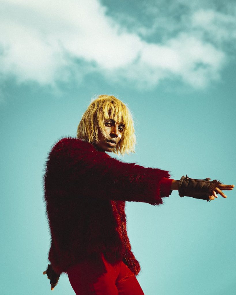 Kabeaushé, a Black person with a yellow chin-length wig, looks into the camera and stands in front of a turquoise sky with a few clouds. Kabeaushé is wearing a wine-red fur coat, their right arm with a brown fur glove is pointing out of the picture on the right.