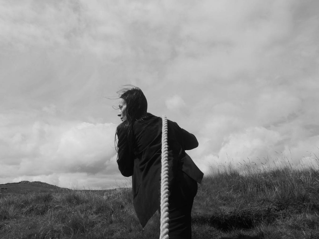 Black and white image of a white woman with long dark hair from behind. Keeley Forsyth is wearing a suit and is pulling a heavy rope over her shoulder, which ends at the bottom of the picture, across a hilly meadow. The sky is overcast.