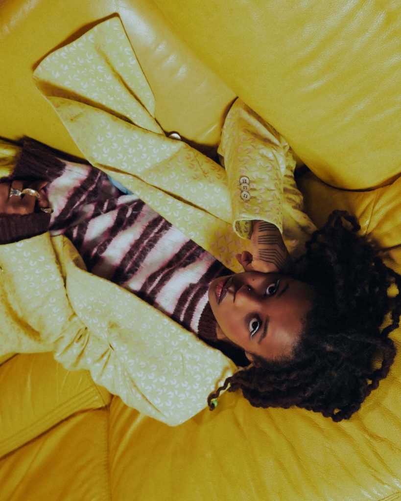 Black, female person with long dreadlocks photographed from above, lying on a yellow leather couch. Lady Donli is wearing a yellow, patterned blazer and a white and purple striped shirt underneath. She rests her face on her right fist and looks into the camera.