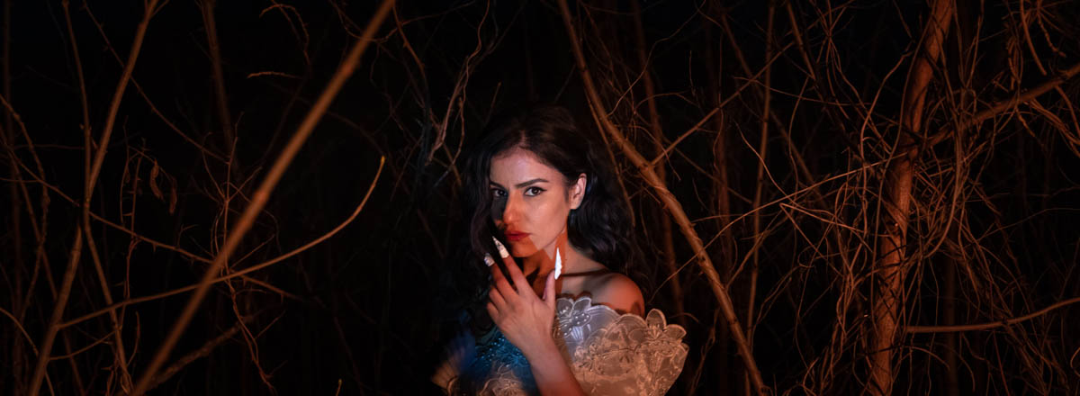 Panoramic shot of a woman with dark hair, a white lace top that leaves her shoulders bare, standing in the dark surrounded by thin twigs and branches of leafless trees. Pari Eskandari looks into the camera, wearing long, white, pointed fingernails and holding her hand up as if to show them.