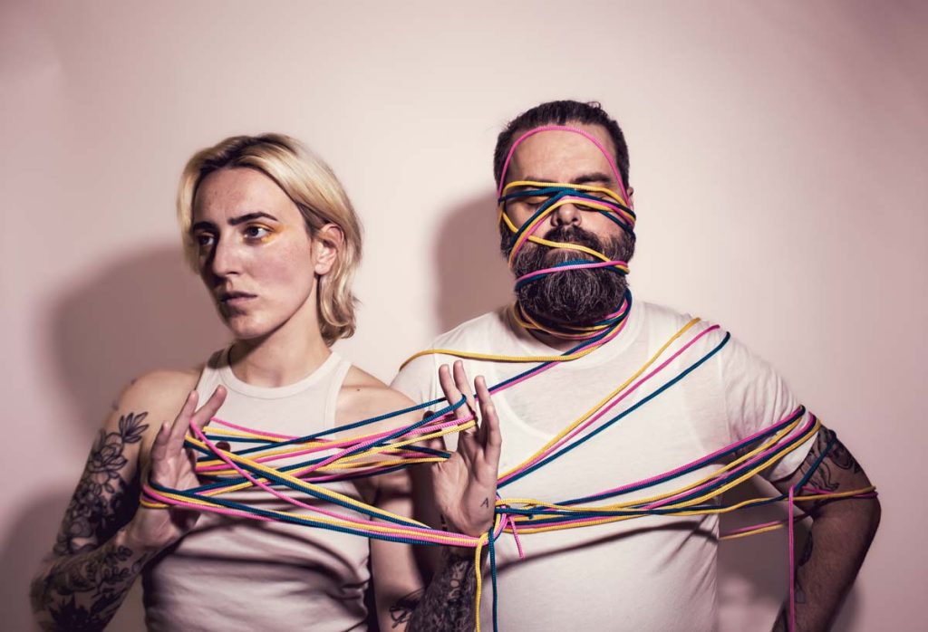 The duo Pillow Fite wear white shirts and stand in front of a white background. Pink, yellow and blue strings are wrapped around the hands of Art Ross and the face of Aaron Green, connecting the two in this way. Art Ross has tucked the shoulder-length blonde hair behind their ears. Aaron Green's eyes are closed by the laces and he wears a full black and grey mottled beard.
