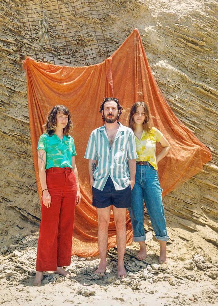 Three people, two women and a man in the centre, stand next to each other in front of an orange cloth hung outside in front of a rock. The members of the band Rasco are all barefoot. He has a full beard, wears a short shirt and shorts with his hands in his pockets. The one on the left with shoulder-length curly hair wears long red trousers and a green blouse, the one on the right with long straight hair wears jeans and a yellow blouse. It is sunny.