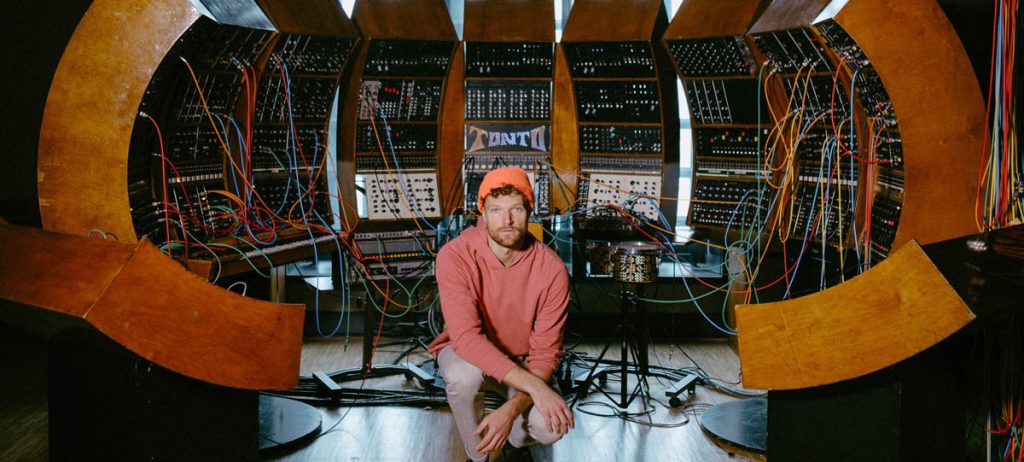 A man in a bright red sweatshirt and neon orange cap squats in the middle in front of a semi-circular wall full of mixing consoles with colourful cables in them. The word 