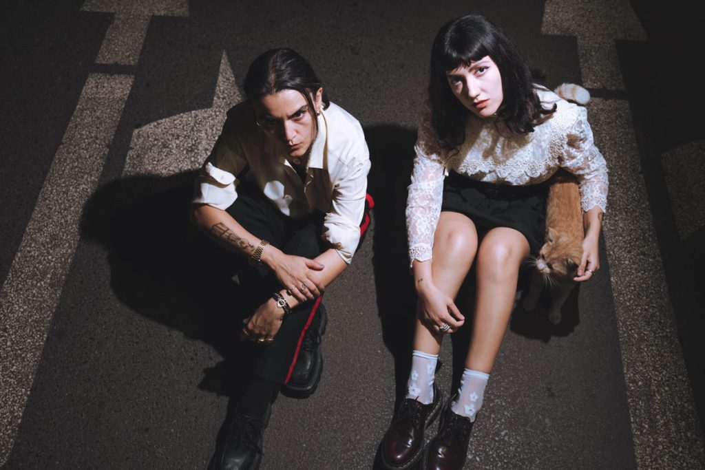 Two female figures with black hair are sitting on a road with arrows pointing forwards and to the right. The band Stereotype was photographed from above. The one on the right is wearing long black trousers and a white shirt with the sleeves rolled up, while the one on the left is wearing a white frilled blouse and a short black skirt. Next to her is a red cat, around which she has her arm.