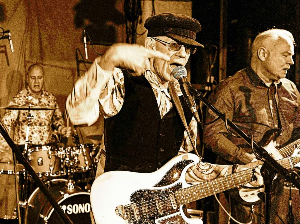 Monochrome picture showing three older men on a stage. The one in front, wearing a cap, darkened glasses and a black waistcoat over a strikingly patterned shirt, sings into a microphone, plays the guitar and gesticulates. The drummer and another guitarist can be seen in the background.