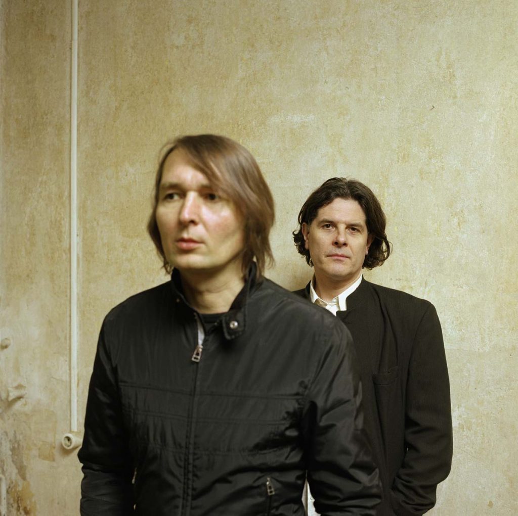Two men dressed in black stand one behind the other in front of a beige-coloured wall. The band Tarwater. The focus is on the man in the back, who is also looking at the camera. The man in front is slightly out of focus and looking to the left.
