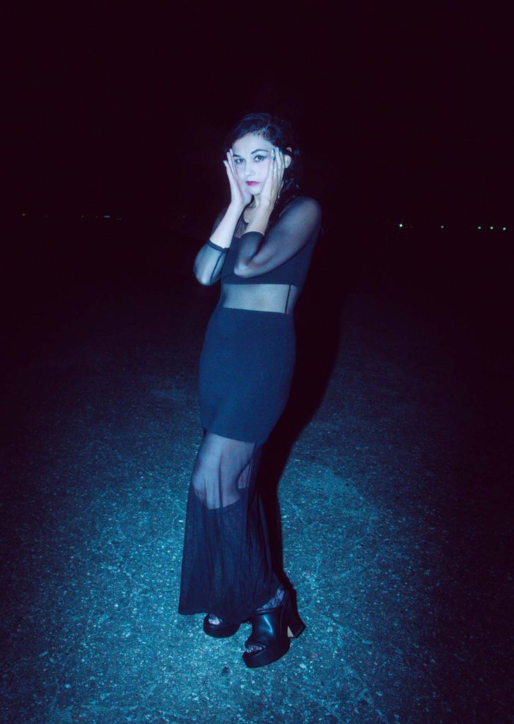A woman stands in the dark in a long black dress that is partly transparent. Light falls only on VV. She puts her palms to her cheeks and looks into the camera.