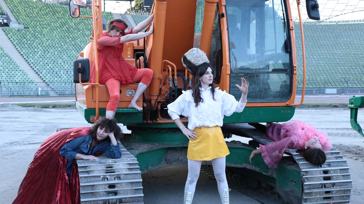 The four members of "What Are People For?", three women and one man, are standing, sitting or lying down and leaning against an orange-coloured excavator standing in a stadium. The green rows of seats can be seen in the background. The musicians are wearing eye-catching, colourful clothing.