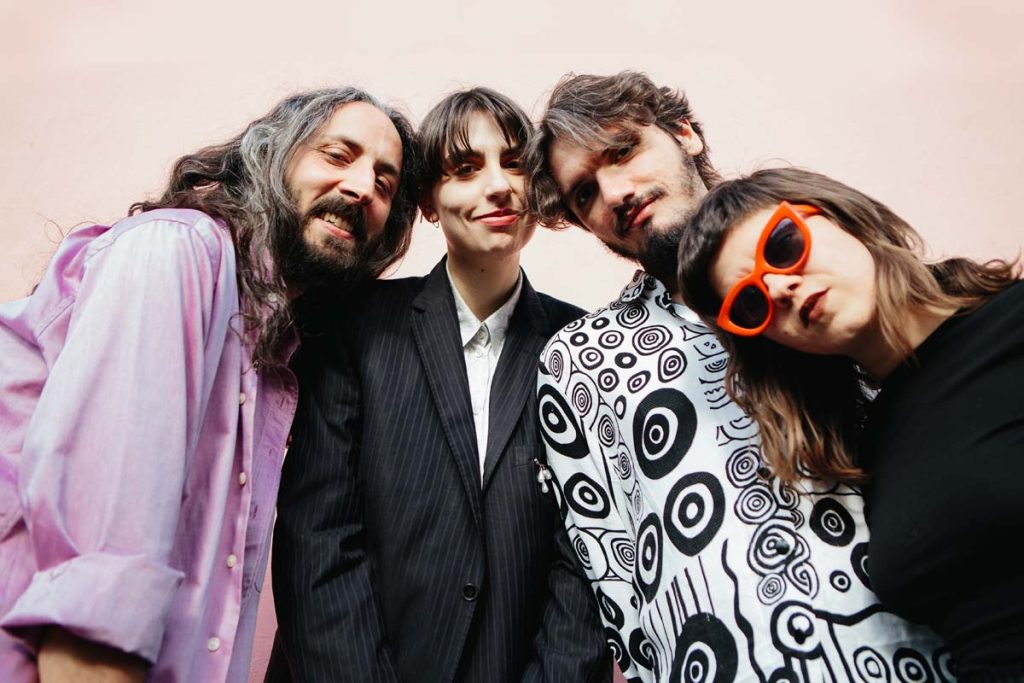 The four members of the band ZIMBRU lean close to each other. The man on the left has long black-grey mottled hair, a full beard and is laughing. Next to him is a female figure with short dark hair, wearing a pinstripe suit. Next to her is a man with brown hair, a beard and a white shirt with black circular patterns. The woman on the far right is wearing large red sunglasses, long brown hair with short fringes and a furrowed brow.