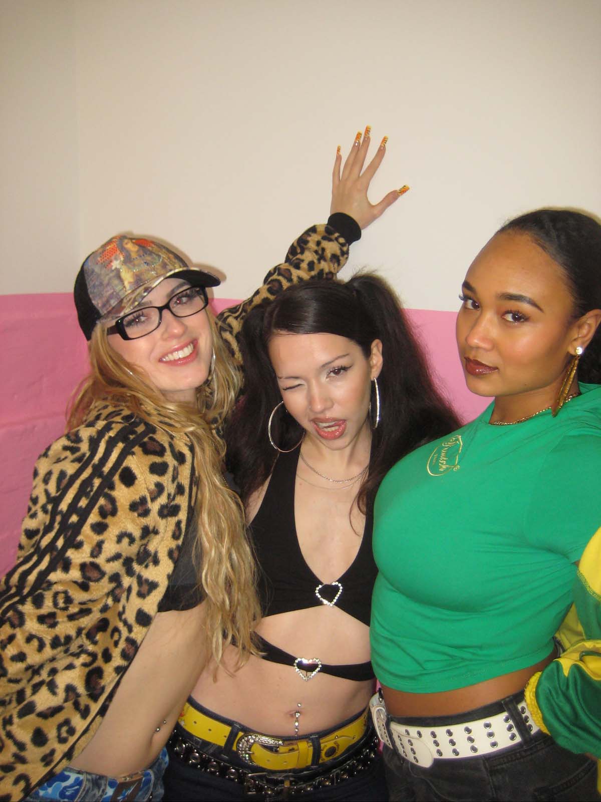 Three female rappers from the crew bangerfabrique stand close together and look into the camera. The white person on the left has long blonde hair, wears a black crop top, baseball cap, leopard tracksuit top, glasses, smiles broadly and rests her left hand on the wall with long fingernails. The one in the centre has one eye closed, wears her dark brown hair tied into two plaits, large silver hoop earrings and a black crop top. The black person on the right has her black hair tied back tightly and is wearing a green T-shirt. They are all wearing trousers and wide 90s-style belts.