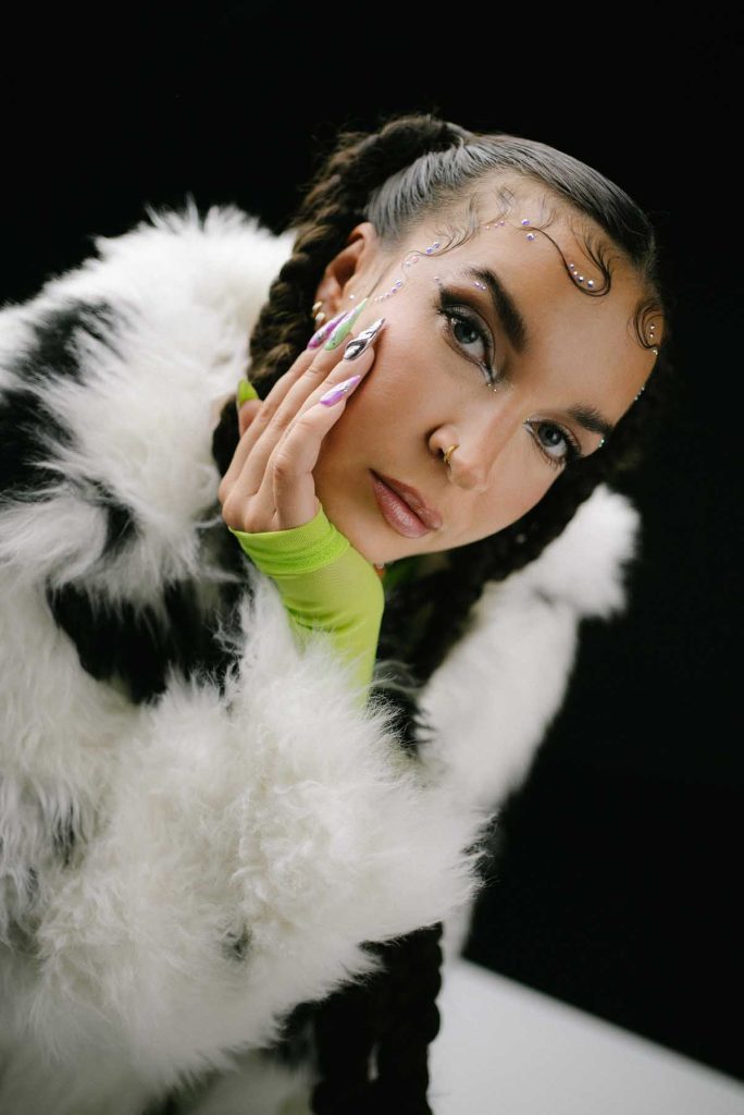 Close-up of a white woman looking into the camera and leaning her head on her hand with long fingernails of different colours. futurebae is wearing a black and white fake fur jacket over a poison green top. Her brown hair is tied back in braids. Her baby hair is fixed to her forehead and glittering stones are decoratively glued on.
