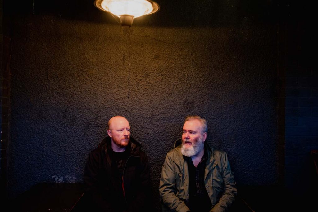 The two members of the band Arab Strap are sitting in front of a dark wall, the scene is lit from above by an old ceiling lamp. The two are only visible in the lower half of the picture. The man on the left is wearing black, is bald, has a red beard and looks almost sadly out of the picture to the bottom right. The one on the right, wearing an olive green jacket, has thinning hair and a full grey beard, looks past the other to the left.