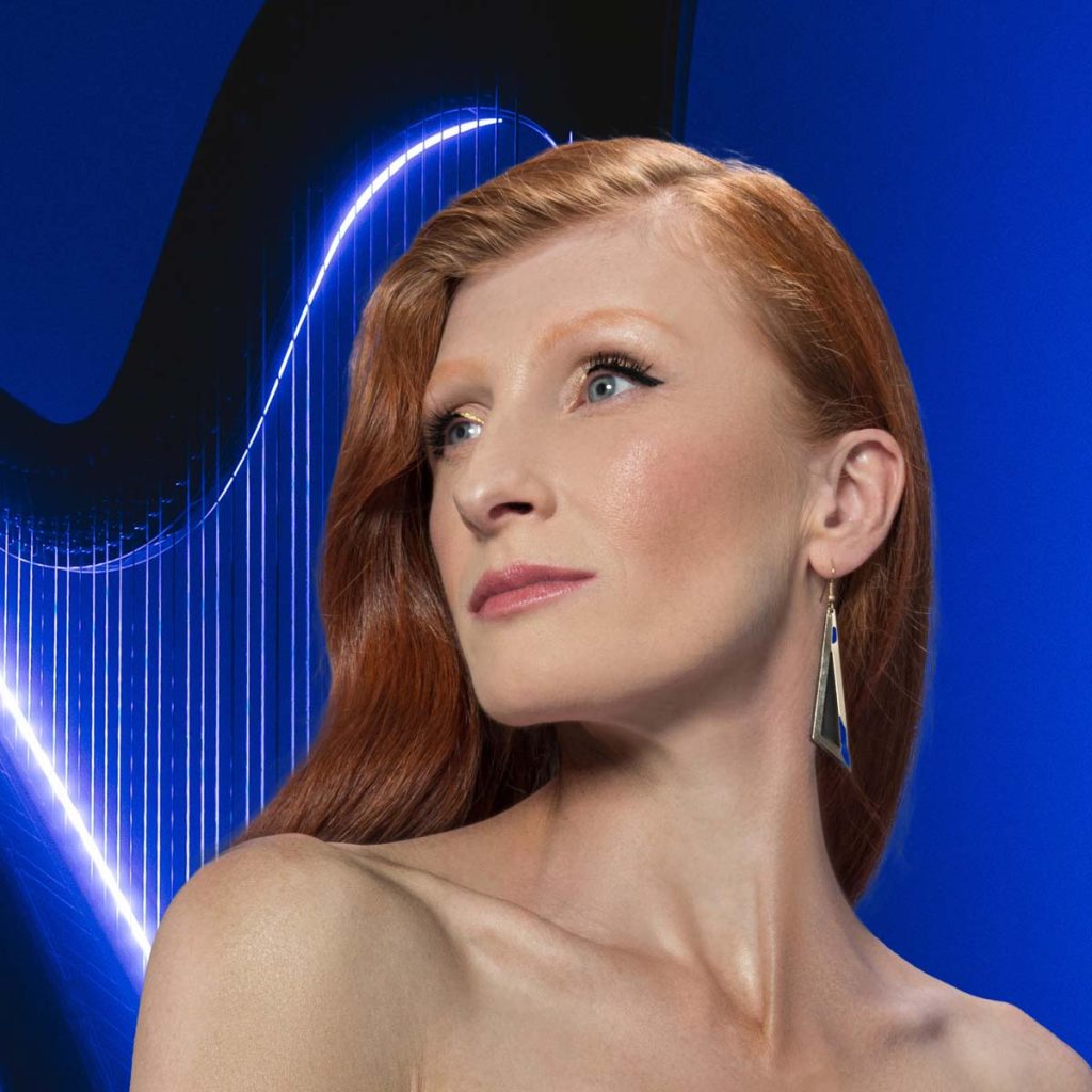 Close-up portrait of a white woman with long red hair. Martina Stock is looking to the left out of the picture. The cropped section only shows her bare shoulders. She is wearing light make-up and a long earring. An illuminated harp can be seen behind her against a dark blue background.
